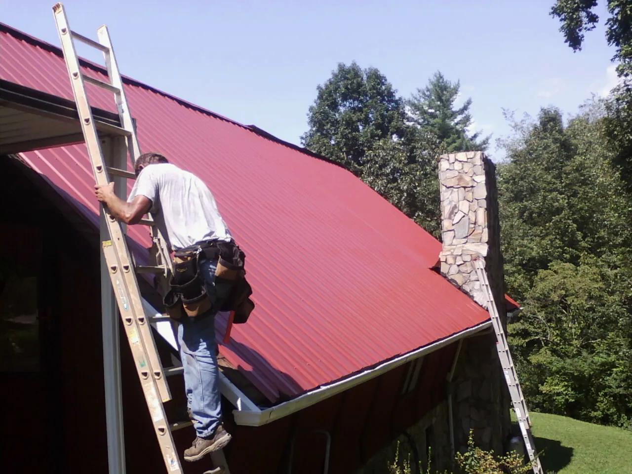 Roofer on a ladder beside a red metal roof and stone chimney — Soffit & Fascia Repair by Roofing Brothers in Waseca